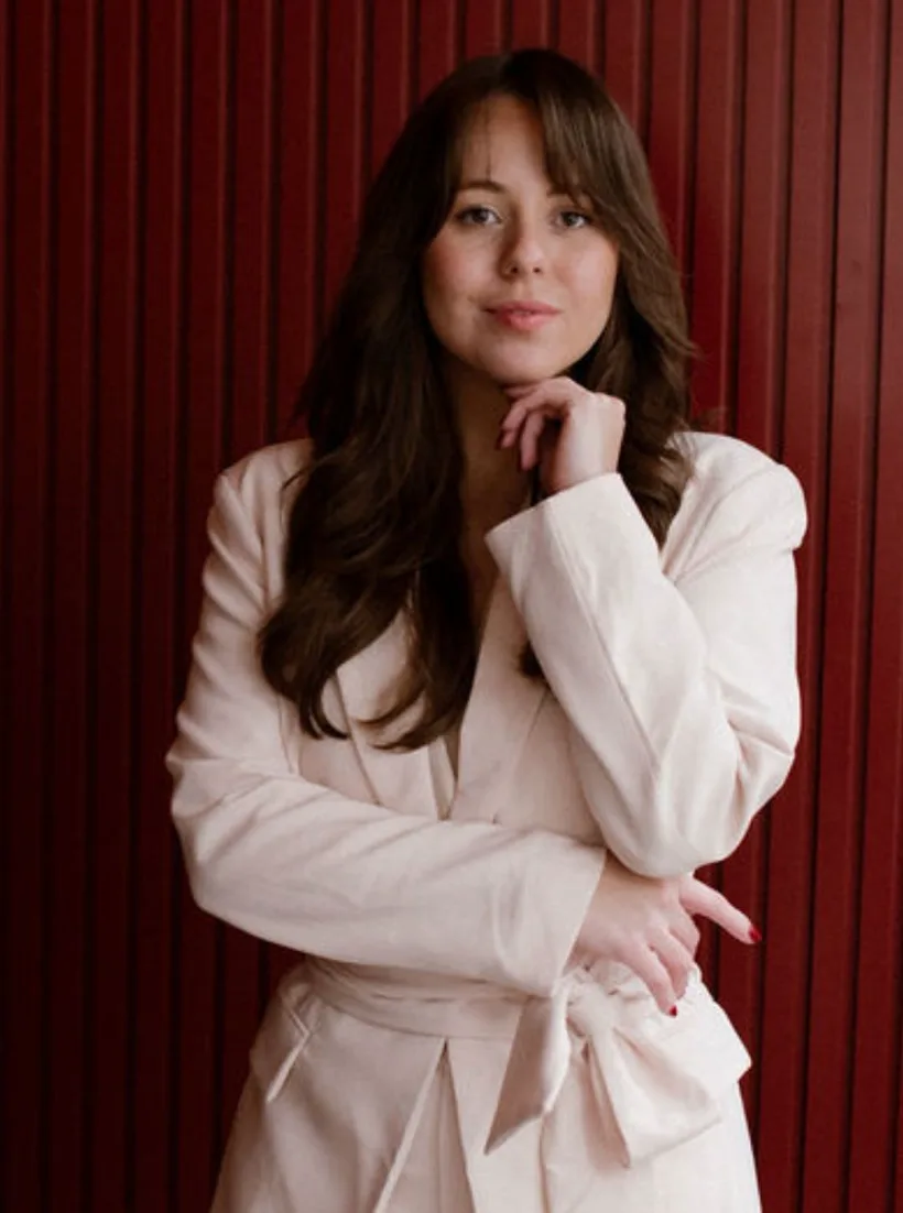 Thessa in a cream blazer against a deep red wall, thinking